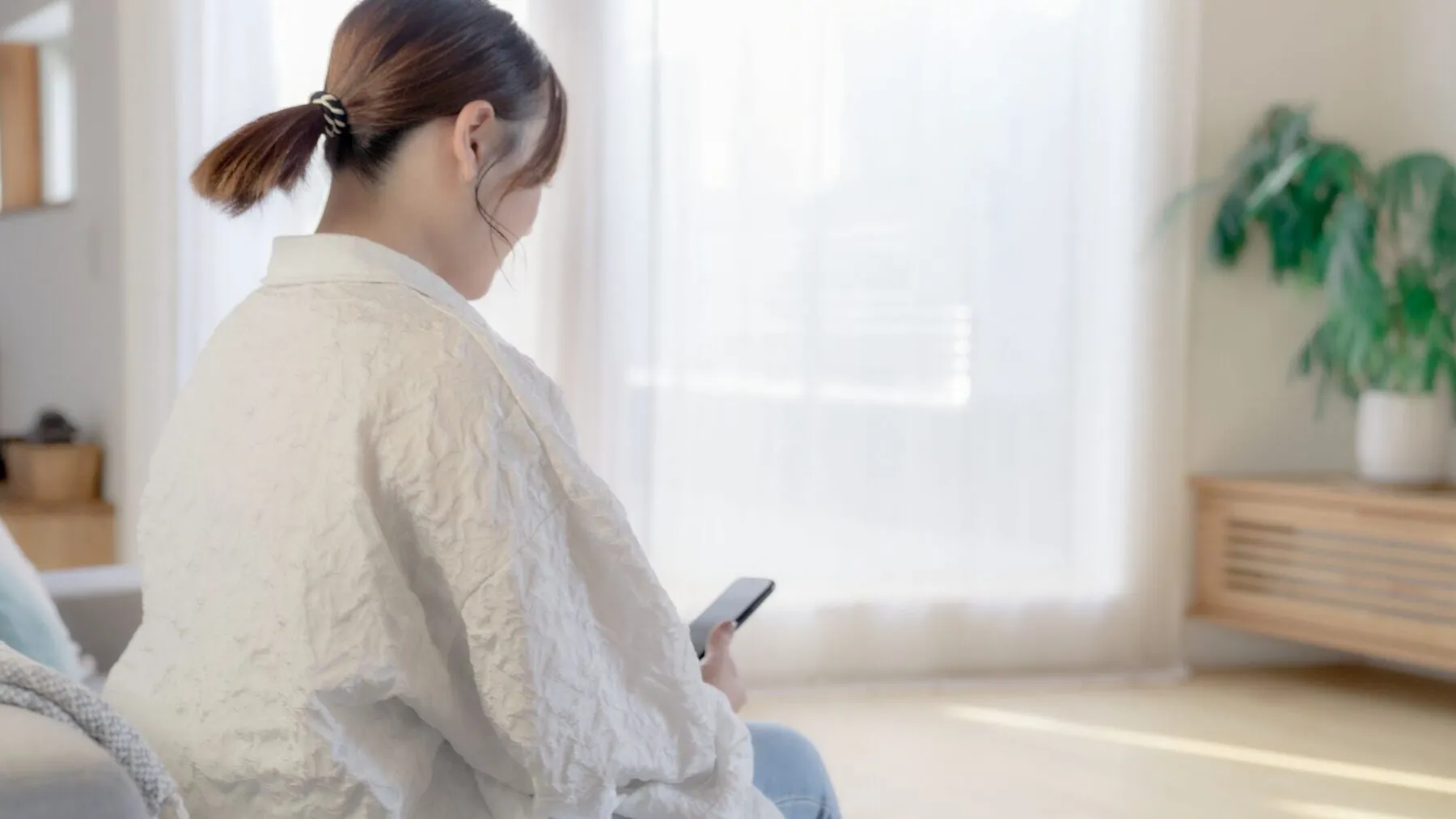 窓辺でスマートフォンを見る女性の後ろ姿